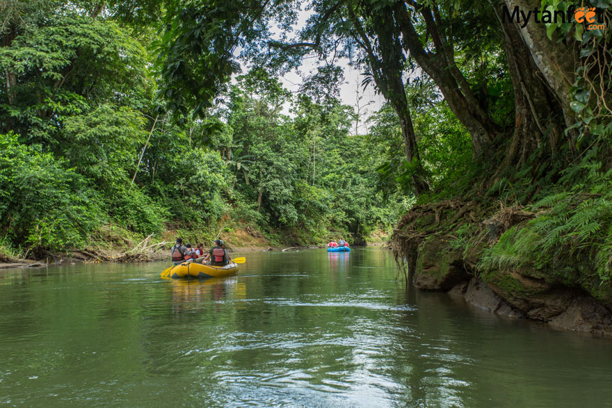 Photo of a river surrounded by green forest, there is a yellow raft with people sitting in it, in the background is a blue raft with people 