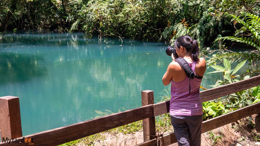 blue lagoon. Photo of a woman of Asian descent holding a camera, wearing a purple tank top looking at a blue lagoon of water 