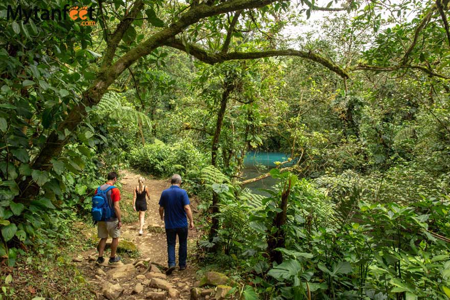 trails. Photo of two men and one woman walking on a natural path surrounded by green rainforest, with a blue river in the distance 