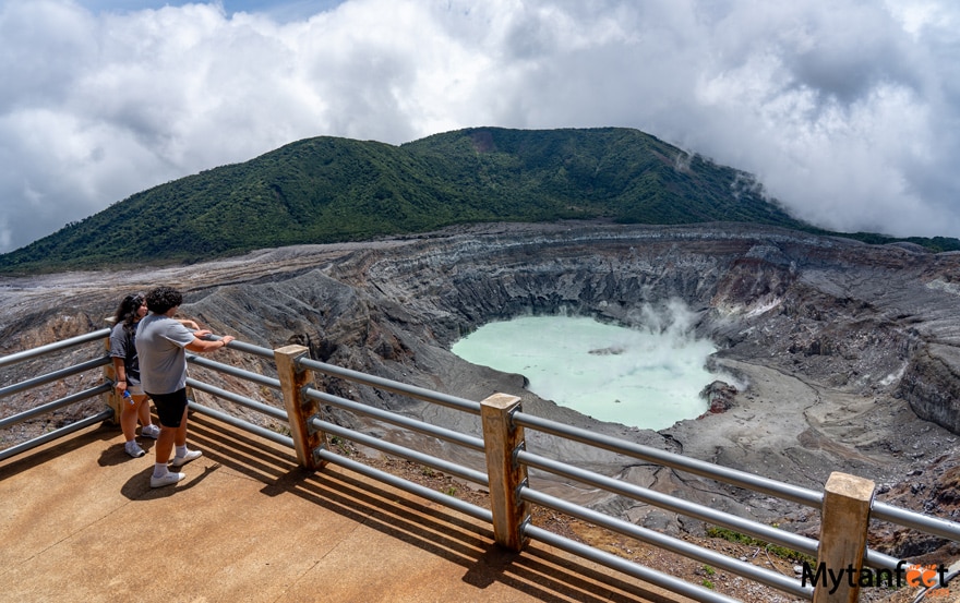 driving from san jose to la fortuna stop at poas volcano national park. A man and woman are standing on an observation platform with a guard rail, looking at a large light blue volcano crater lake that is slightly smoking 