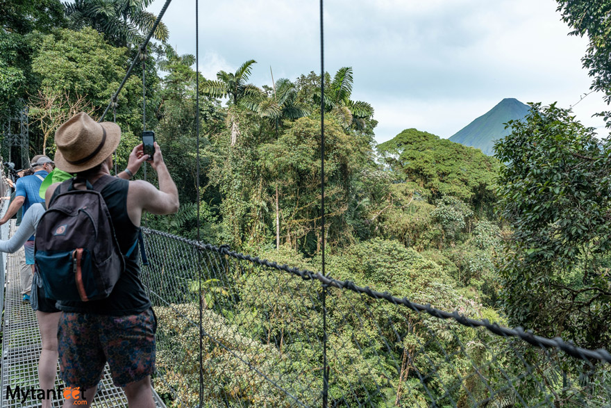 A man in hiking gear is on a hanging bridge in the rainforest, taking a picture with his cell phone of Arenal Volcano in the distance 