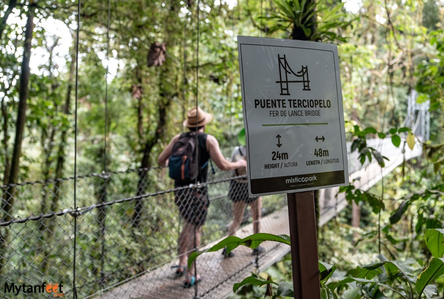 A man is walking on a hanging bridge and the focus is of the sign in front of the bridge saying puente tercipielo, 24 meters in height and 48 meters in length 