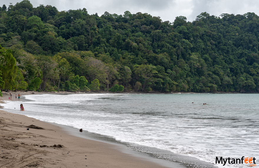 costa rica in november playa herradura. View of a beach with cloudy gray skies, green rainforest and greyish water with a few people swimming in the water, and a few people on shore 