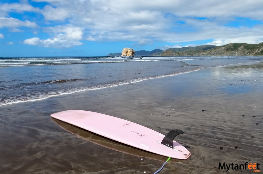 Photo of a pink longboard surfboard, fin up on the shore, with a large rock in the distance behind the beach 