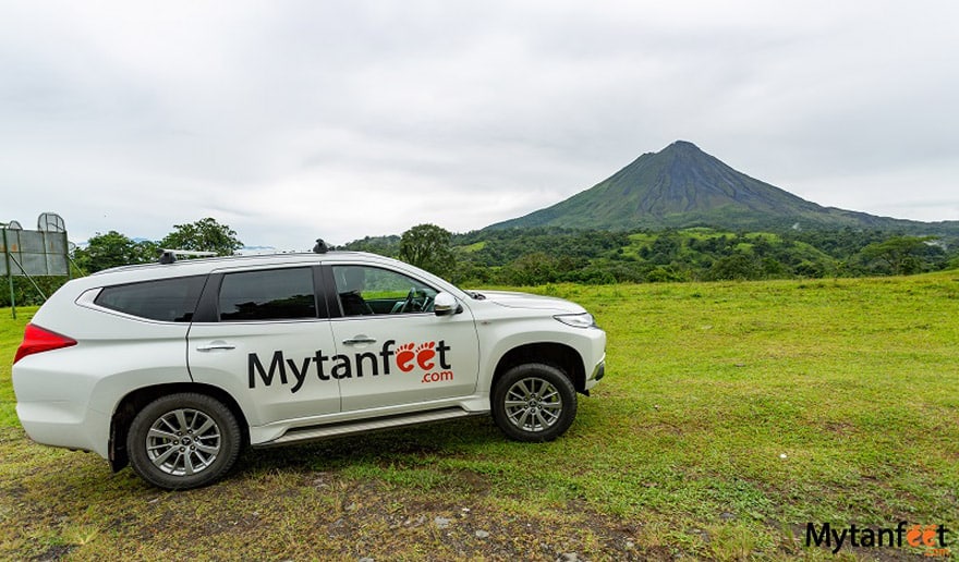 Mytanfeet Costa Rica car rental discount. A white SUV with the logo "MYTANFEET" is parked in on a grassy field, with a volcano in the background in Costa Rica
