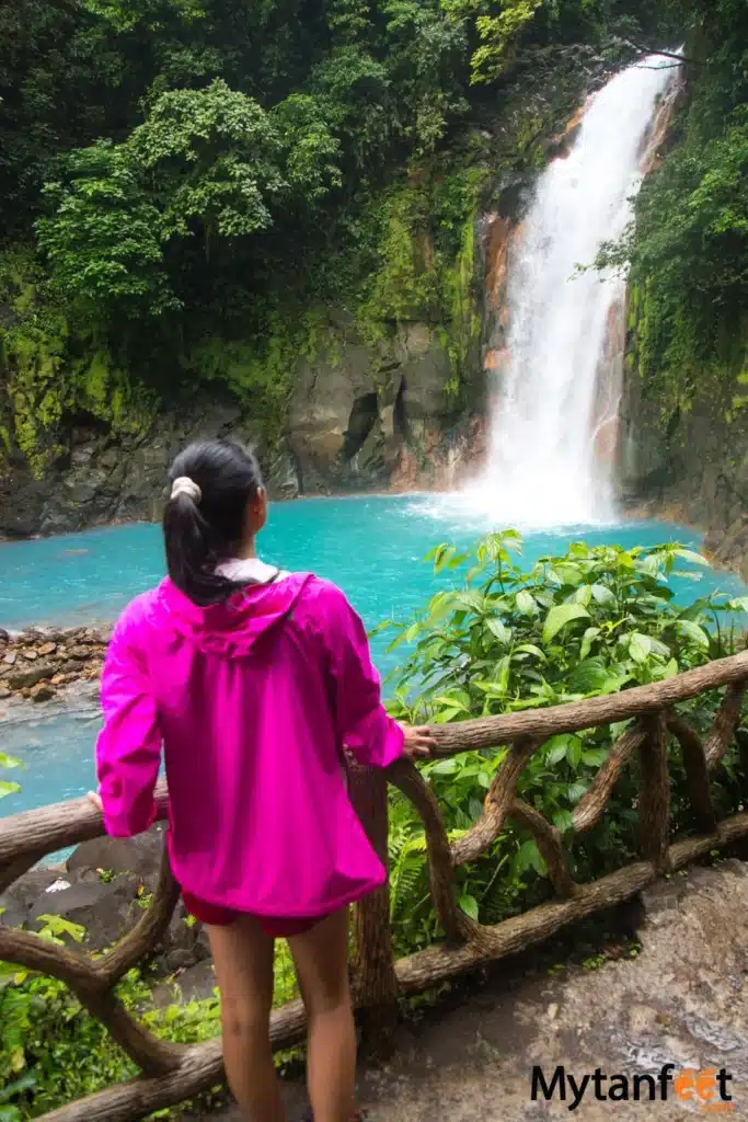 rio celeste waterfall in tenorio volcano national park - an asian woman wearing a pink rain jacket is looking at the waterfall with her back to the camera