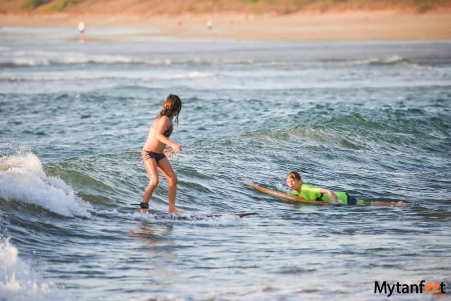 Things to do in Tamarindo with kids - surfing at the beach. A young girl is surfing on a wave and a young boy is paddling out looking at her