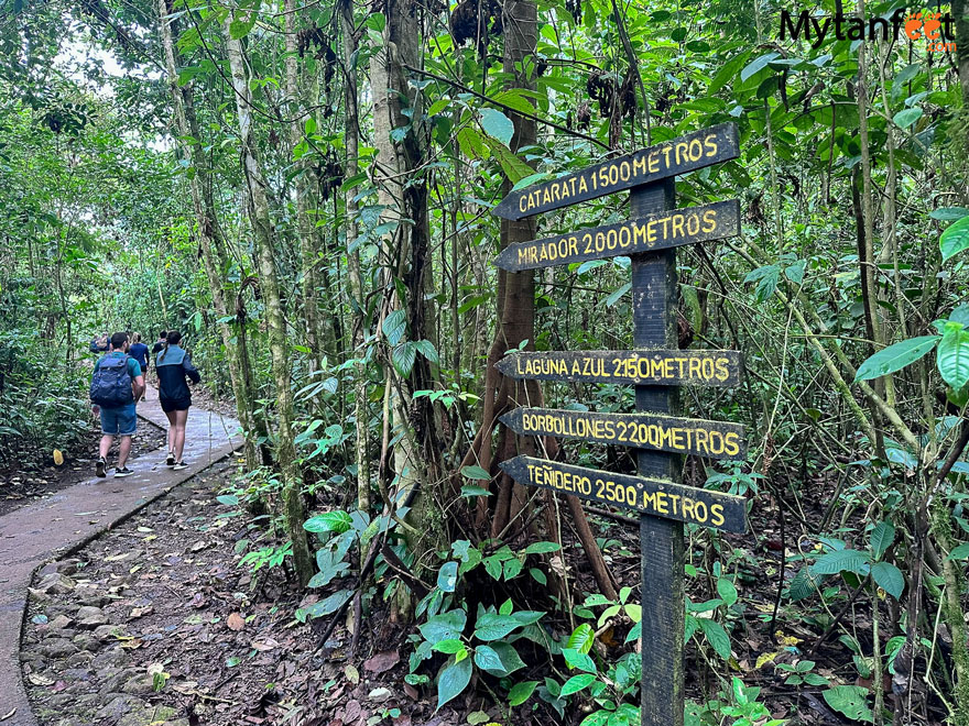 Trail in Tenorio Volcano National Park with a sign saying how far each view point is. Two adults in hiking gear are walking on a concrete path to the left of the sign, inside a rainforest in COsta Rica 