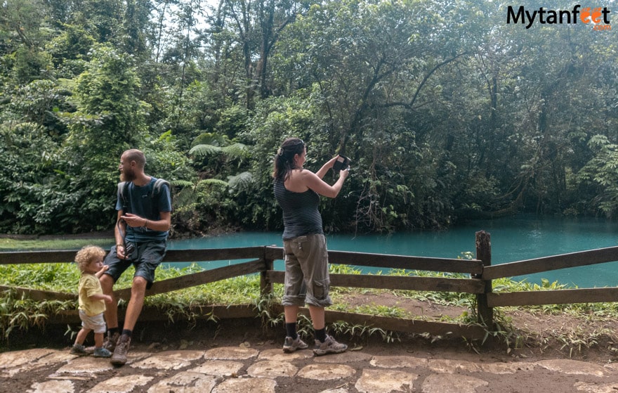 Rio Celeste with kids. A mom is taking a photo on her cell phone of a sky blue river, dad is sitting on the hand rail and a young boy with curly blonde hair is looking at dad 