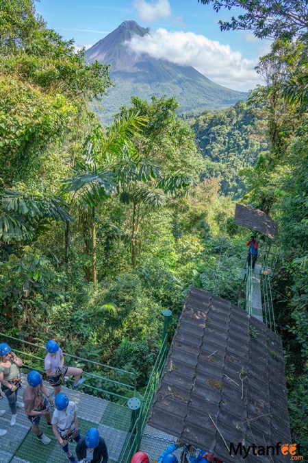 Photo of a group of people dressed in ziplining equipment and gear, standing on a platform waiting to go zipline their first cable, a volcano and rainforest are in the back drop at Sky Adventures in La Fortuna 