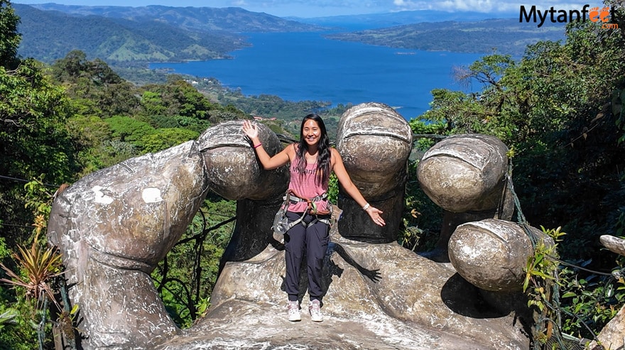 An Asian woman wearing a harness and ziplining gear is standing on a big hand structure, with a lake in the background, posing and smiling at SKy Adventurse in la Fortuna, Costa Rica 