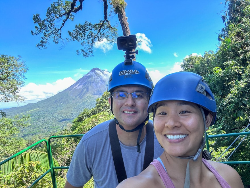Two adults wearing helmets are smiling and posing, taking a selfie during a ziplining tour in la Fortuna. Arenal Volcano and the rainforest are behind them. 