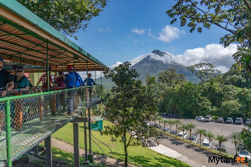 Sky Adventures in La Fortuna - a group of adults is waiting on a platform, with a volcano in the backdrop and parking lot below them, they are wearing helmets and gear to go ziplining in Costa Rica 