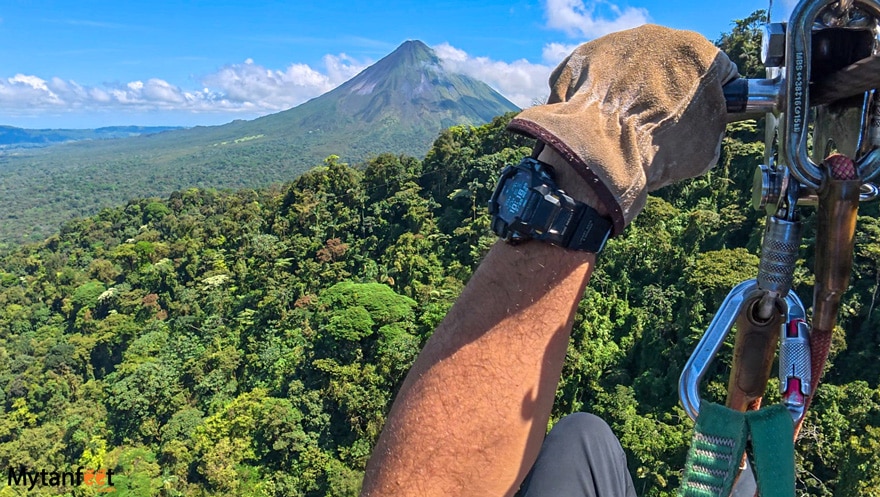 Photo of a man on a ziplining cable, with a volcano and rainforest in front of him, you can only see his left hand wearing a glove holding onto a cable and his harness and gear 