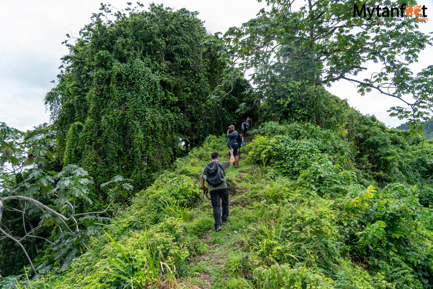 Cocos Island National Park. A man is hiking up a green grassy hill on a dirt trail wearing a backpack and using a hiking pole, another man in front of him is standing, looking at the view