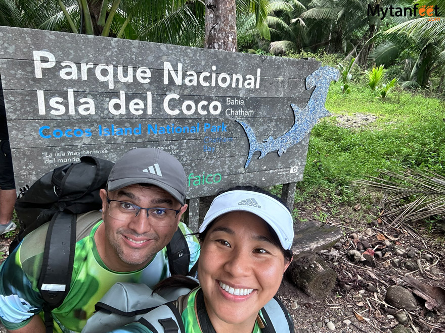 a man and woman both wearing hats and a backpack are taking a selfie, smiling in front of a sign that says Parque Nacional Isla del Coco Bahia Chatham