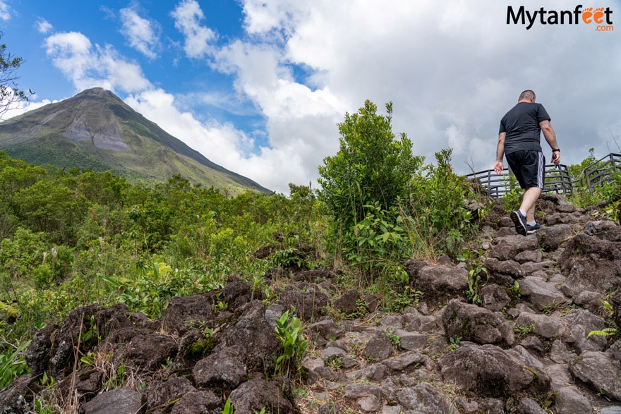 A man is hiking on a trail of lava rocks with a clear volcano to his left inside Arenal Volcano National Park