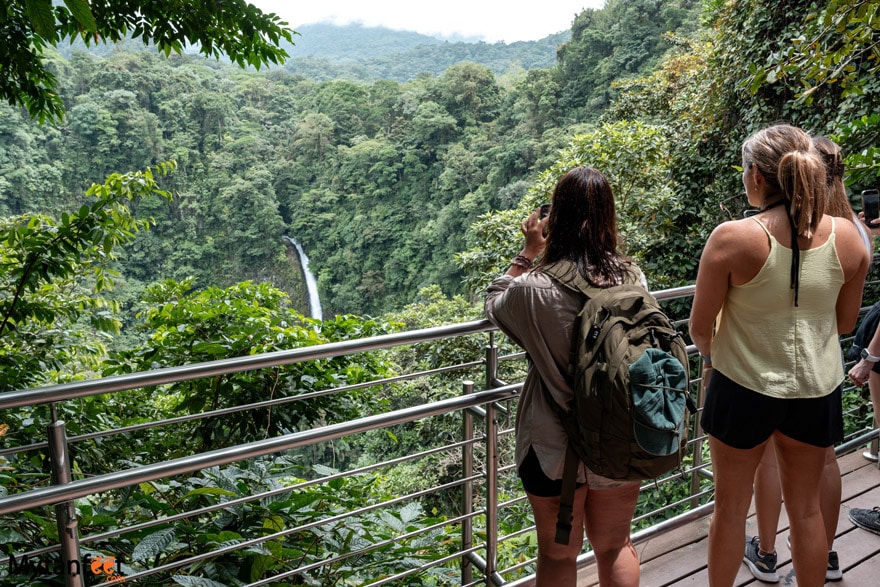 La Fortuna waterfall. Photo of two women taking a photo of a waterfall in the rainforest, standing on a platform with handrails 