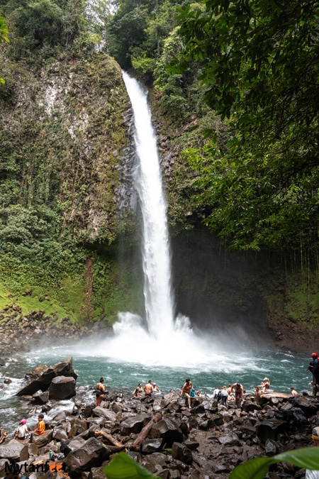 A tall waterfall falling into an emerald green pool, people are sitting on the rocks around the pool of La Fortuna waterfall, Arenal