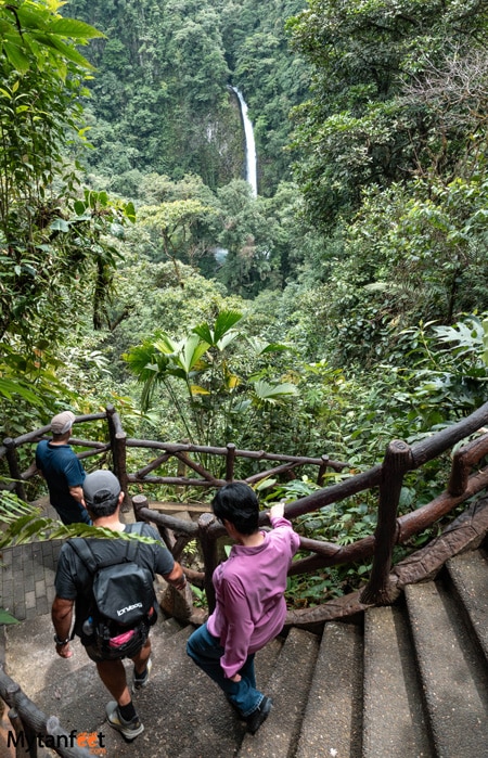 One woman wearing a pink shirt and two men are walking down stairs in the rainforest, with a waterfall in the backdrop in Costa Rica 