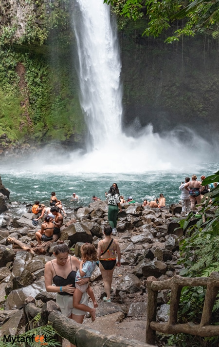 Photo of a big waterfall and emerald colored pool, with a lot of people sitting on the rocks. A mom carrying her child are walking up some stairs away from the La Fortuna waterfall in costa Rica