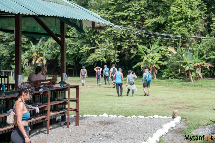  A woman is walking near a building and a group of hikers are in an open field, walking towards the rainforest at Sirena Station in Corcovado National Park 