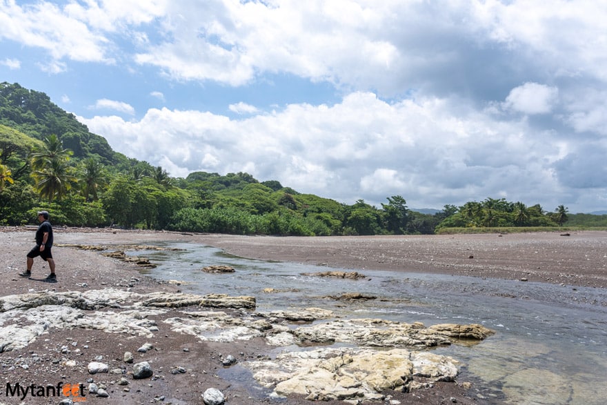 Playa Buena Vista - a man wearing a hat and shorts and a black tshirt is walking on a beach with rocks, with a background of a green forest in Costa Rica 