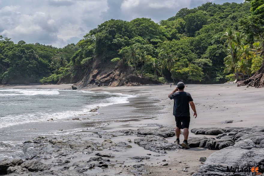 A man dressed in a black shirt and shorts and wearing a baseball hat is walking on the beach, with tan sand and some rocks, with green mountains in the backdrop in Costa Rica