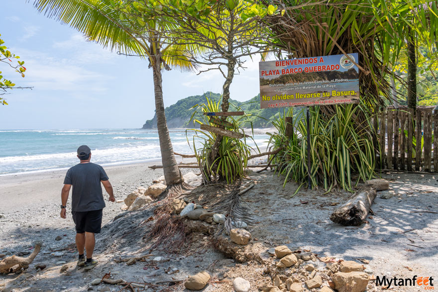 A man wearing shorts and t shirt and hat with shoes is walking onto a beach, with a sign that says bievenidos a playa barco quebrado in Costa Rica