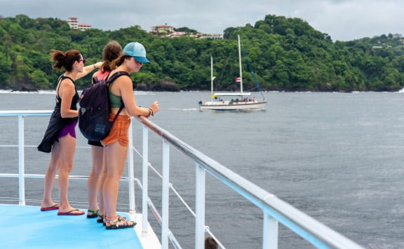 manuel-antonio-catamaran-tour-booking-image manuel antonio catamaran tour three women are standing on a catamaran looking out into the coastline and ocean with a small yacht in the background